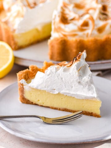 A slice of lemon meringue pie shown on a plate, the whole pie is in the background.