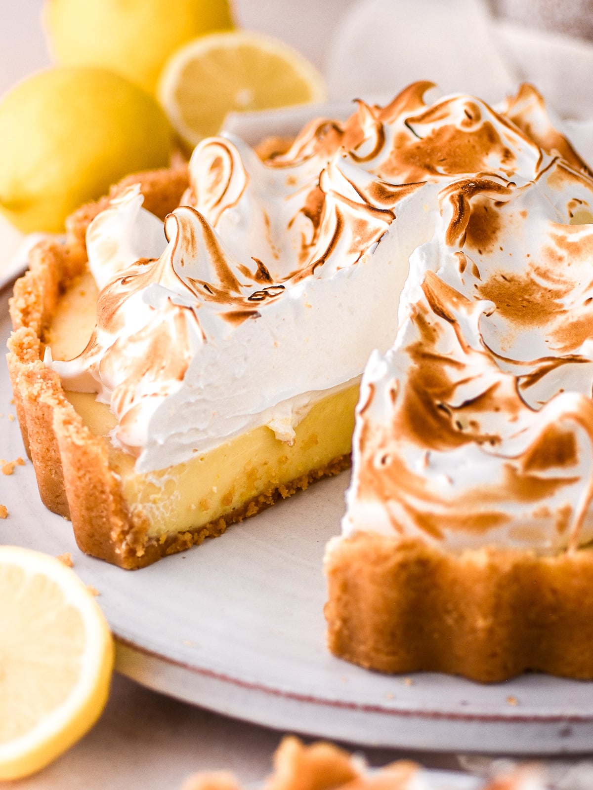 Lemon meringue pie shown on a cake plate, one slice is missing and you can see the layers of biscuit base, lemon filling, and fluffy meringue.