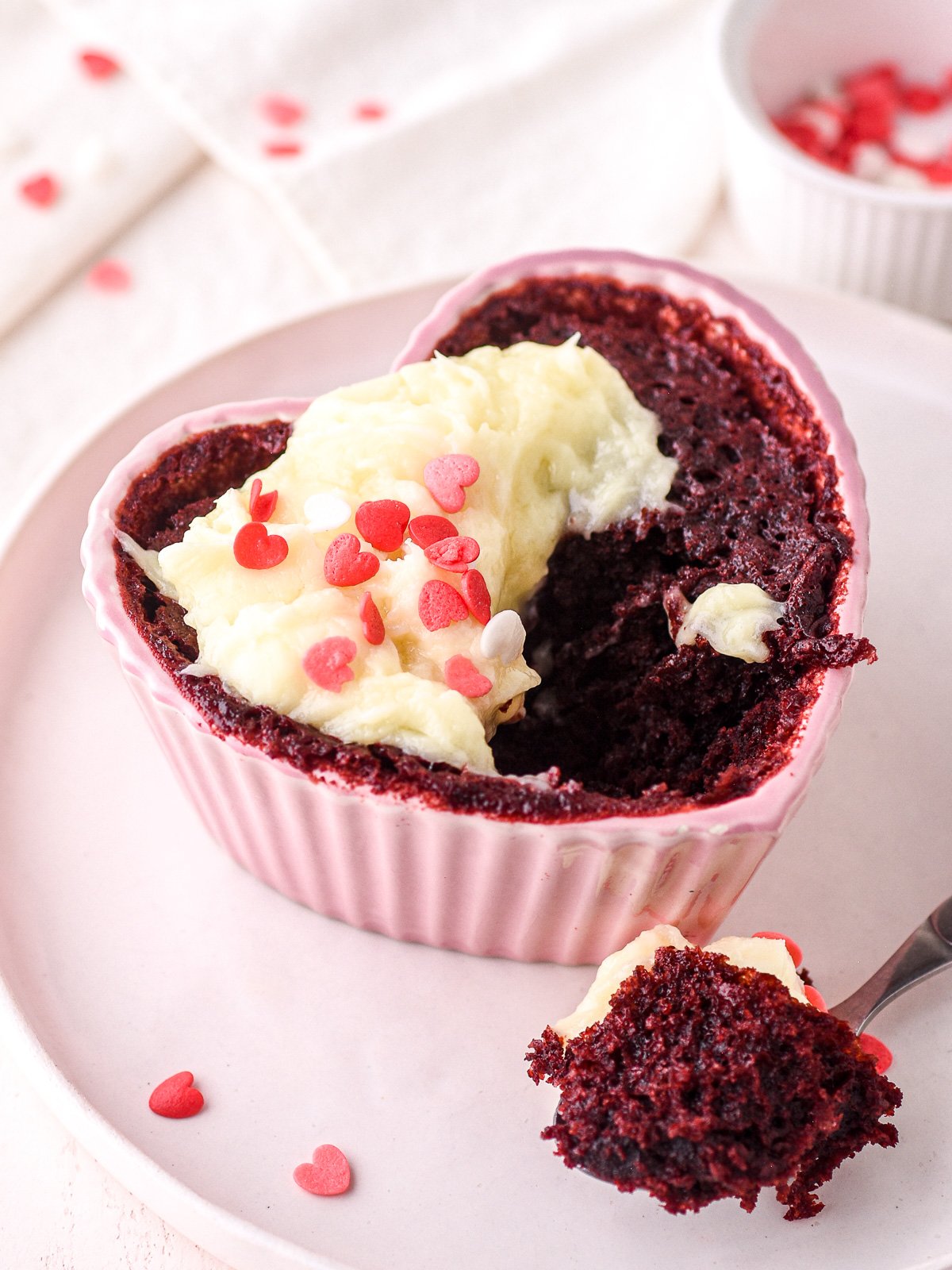 Red Velvet Mug Cake shown in a heart shaped ramekin with a spoon next to it.
