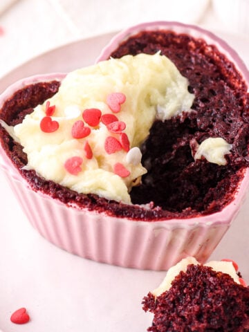 Red velvet mug cake baked into a pink heart shaped ramekin and topped with cream cheese frosting and heart sprinkles.