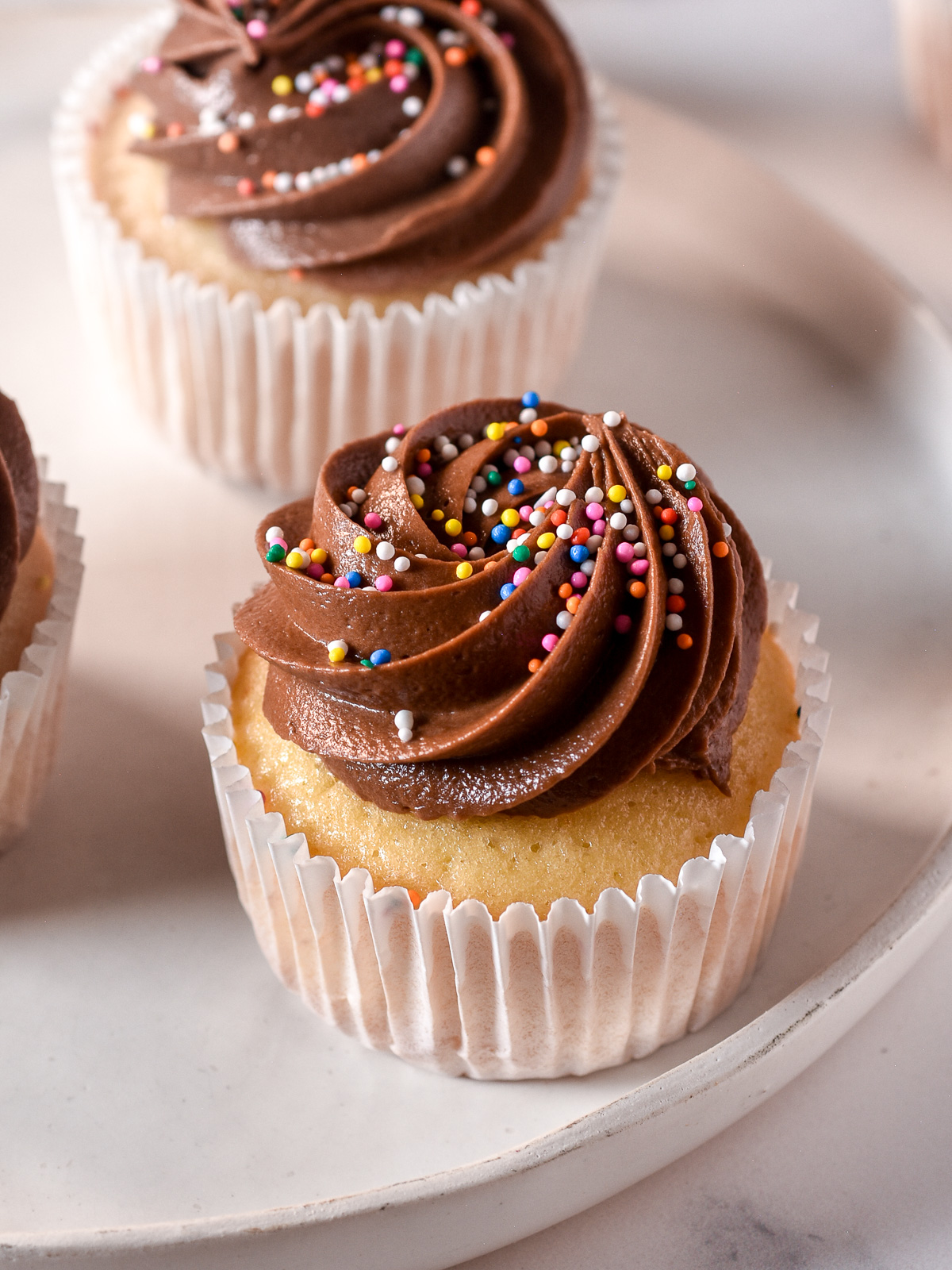 Vanilla chocolate cupcake shown on a white plate. It has a perfect swirl of chocolate frosting that has been topped with sprinkles.