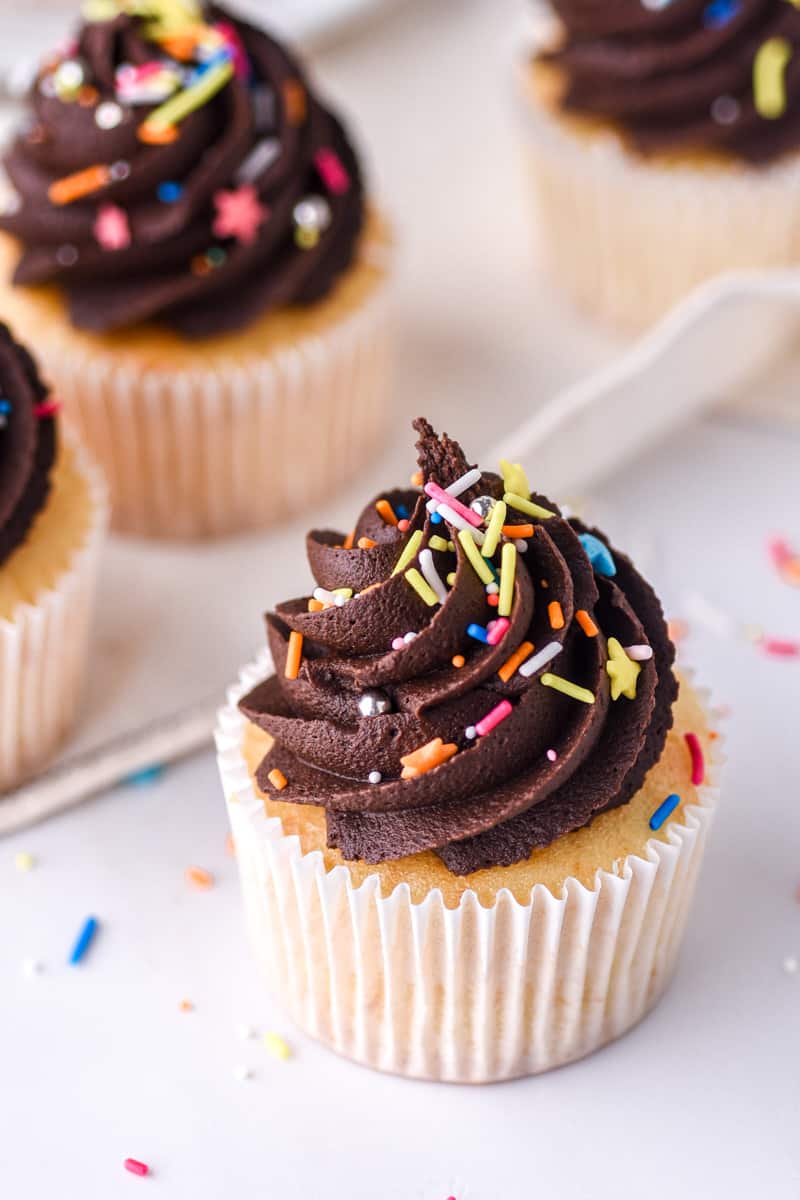 Vanilla Cupcakes with Chocolate Frosting shown decorated with sprinkles on a kitchen counter. 