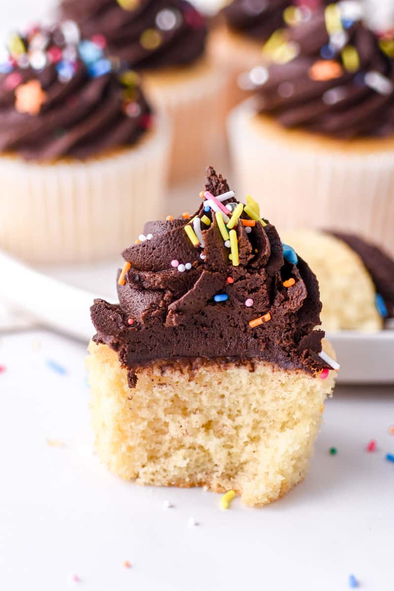 Vanilla Cupcakes with Chocolate Frosting shown on a white kitchen counter, the cupcake in the front has a bite missing so that you can see the fluffy texture of the cupcake. 
