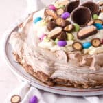 Easter pavlova decorated with different types of easter eggs shown on a serving plate on a pink kitchen counter.