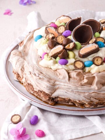Easter pavlova decorated with different types of easter eggs shown on a serving plate on a pink kitchen counter.