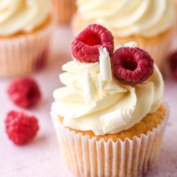 Raspberry and white chocolate cupcakes show on a kitchen counter next to fresh raspberries.