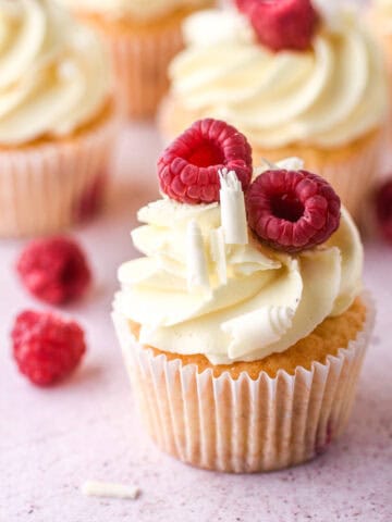 Raspberry and white chocolate cupcakes show on a kitchen counter next to fresh raspberries.