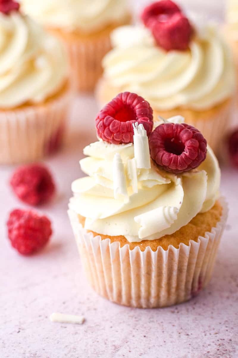 Raspberry and White Chocolate Cupcake shown u close with other cupcakes blurred in the background. 