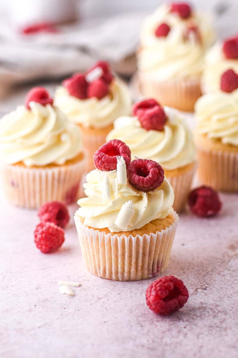 Raspberry and White Chocolate Cupcakes shown on a kitchen counter with fresh raspberries and curls of white chocolate. 