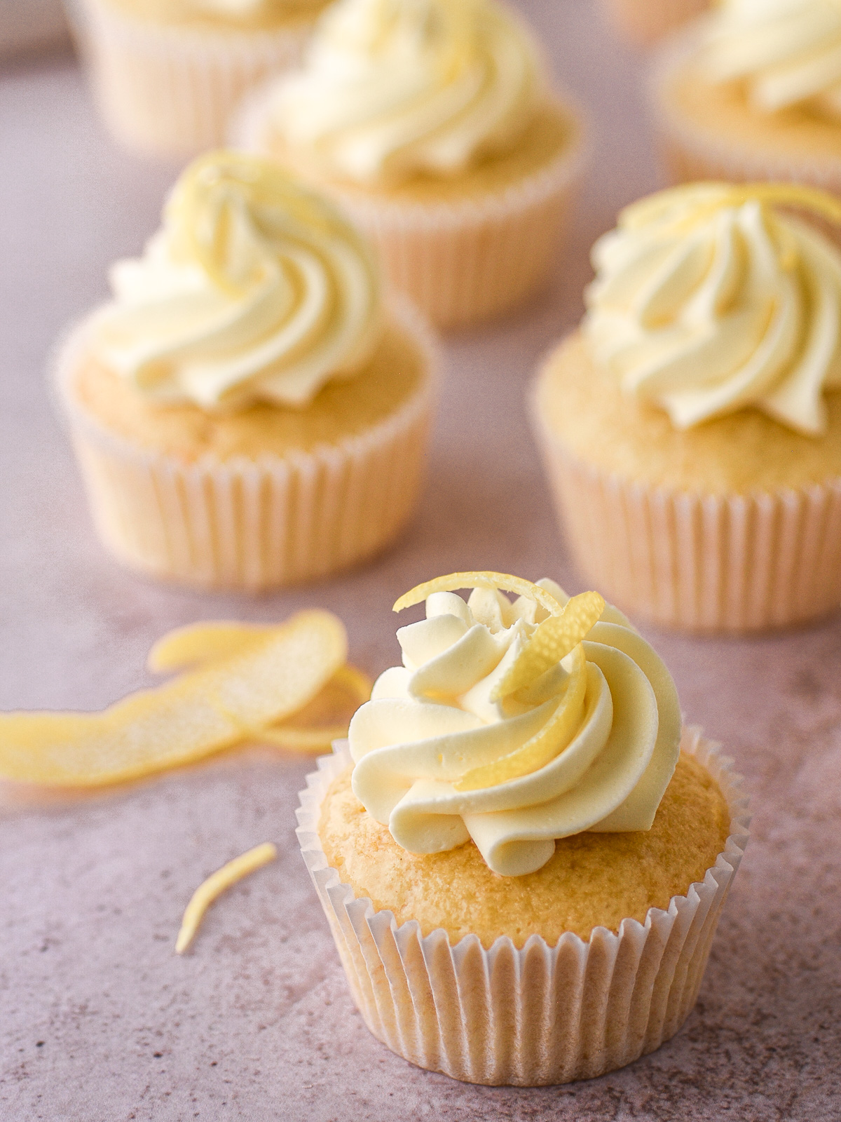Lemon drizzle cupcake decorated with vanilla buttercream and lemon zest shown on a kitchen counter. There are more lemon cupcakes in the background.