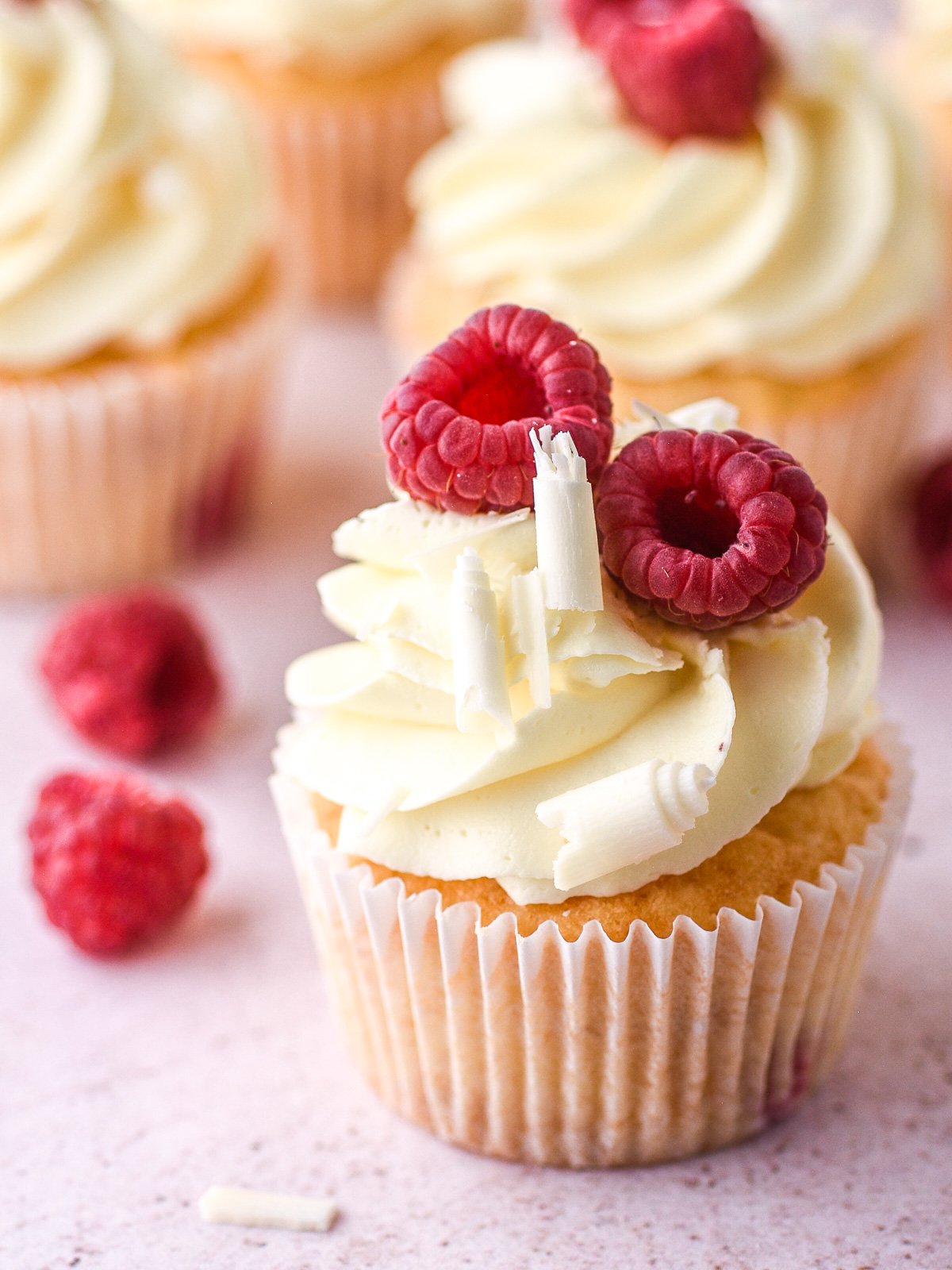 Image shows a raspberry and white chocolate cupcake on a counter, it is decorated with chocolate curls and raspberries.