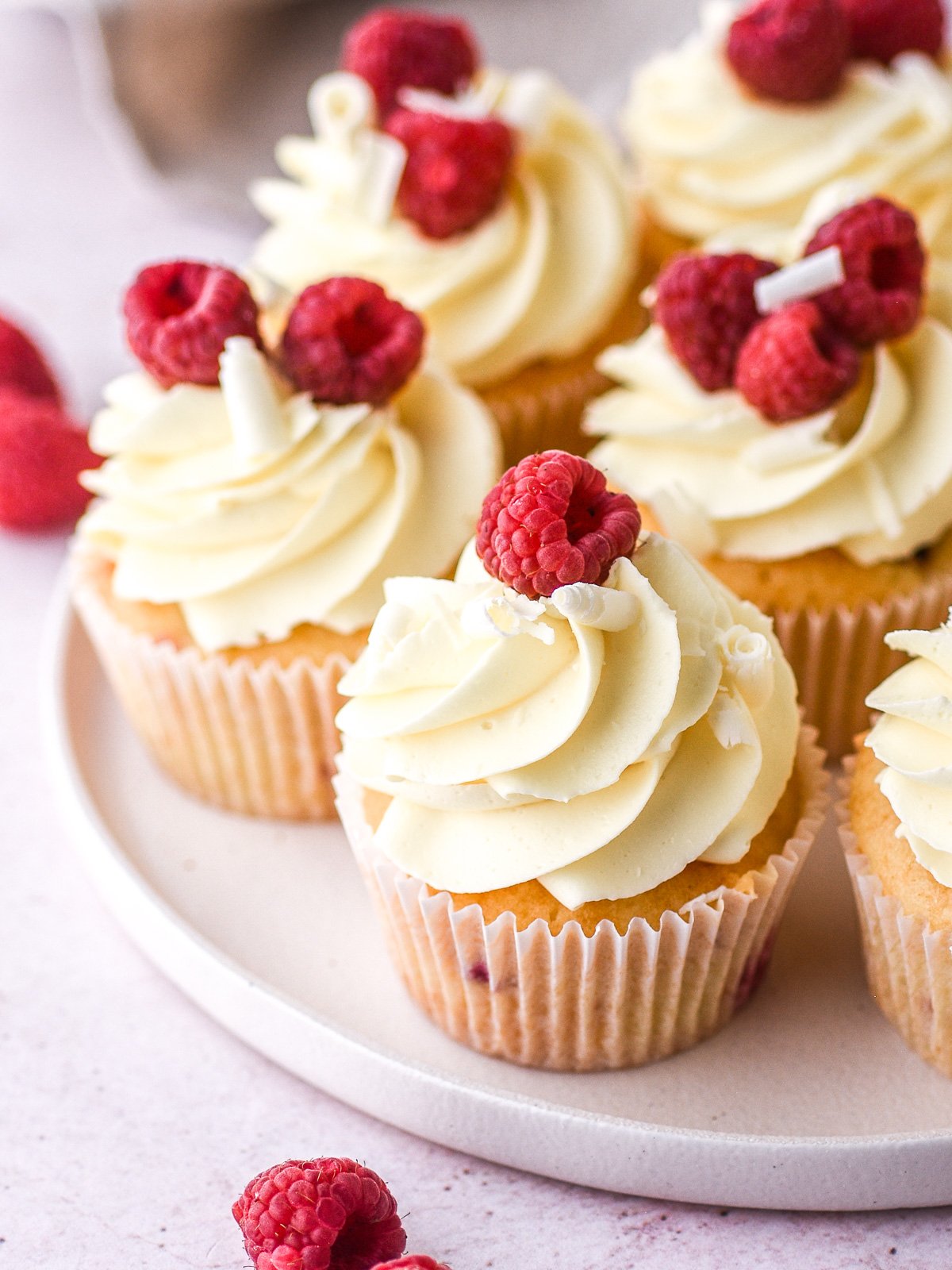 Decorated cupcakes shown on a white plate with fresh raspberries scattered around the plate.