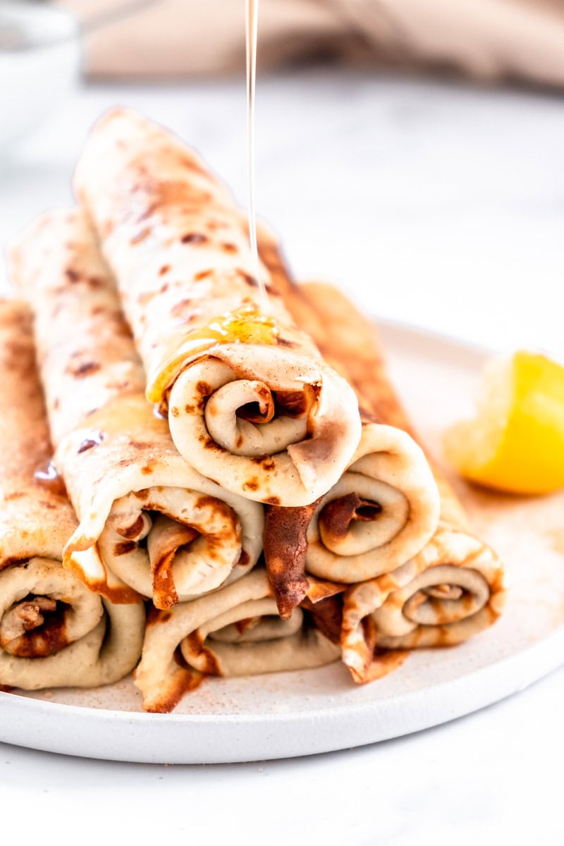 South African Pancakes with Cinnamon Sugar shown stacked on a white plate with honey being drizzled over them.
