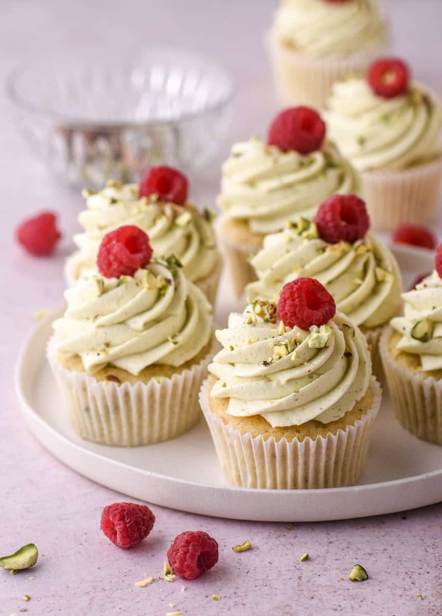 Pistachio Buttercream show piped onto cupcakes. These are cupcakes shown on a plate decorated with pistachio buttercream and raspberries. 