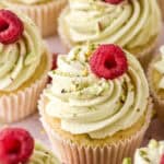 Pistachio Cupcakes shown on a kitchen counter, you can see one cupcake up close that has been decorated with a fresh raspberry and crushed pistachios.
