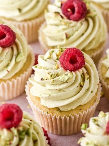Pistachio Cupcakes shown on a kitchen counter, you can see one cupcake up close that has been decorated with a fresh raspberry and crushed pistachios.