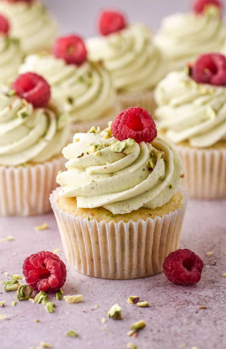 Pistachio Cupcakes shown on a kitchen counter. They have been decorated with raspberries and crushed pistachios.