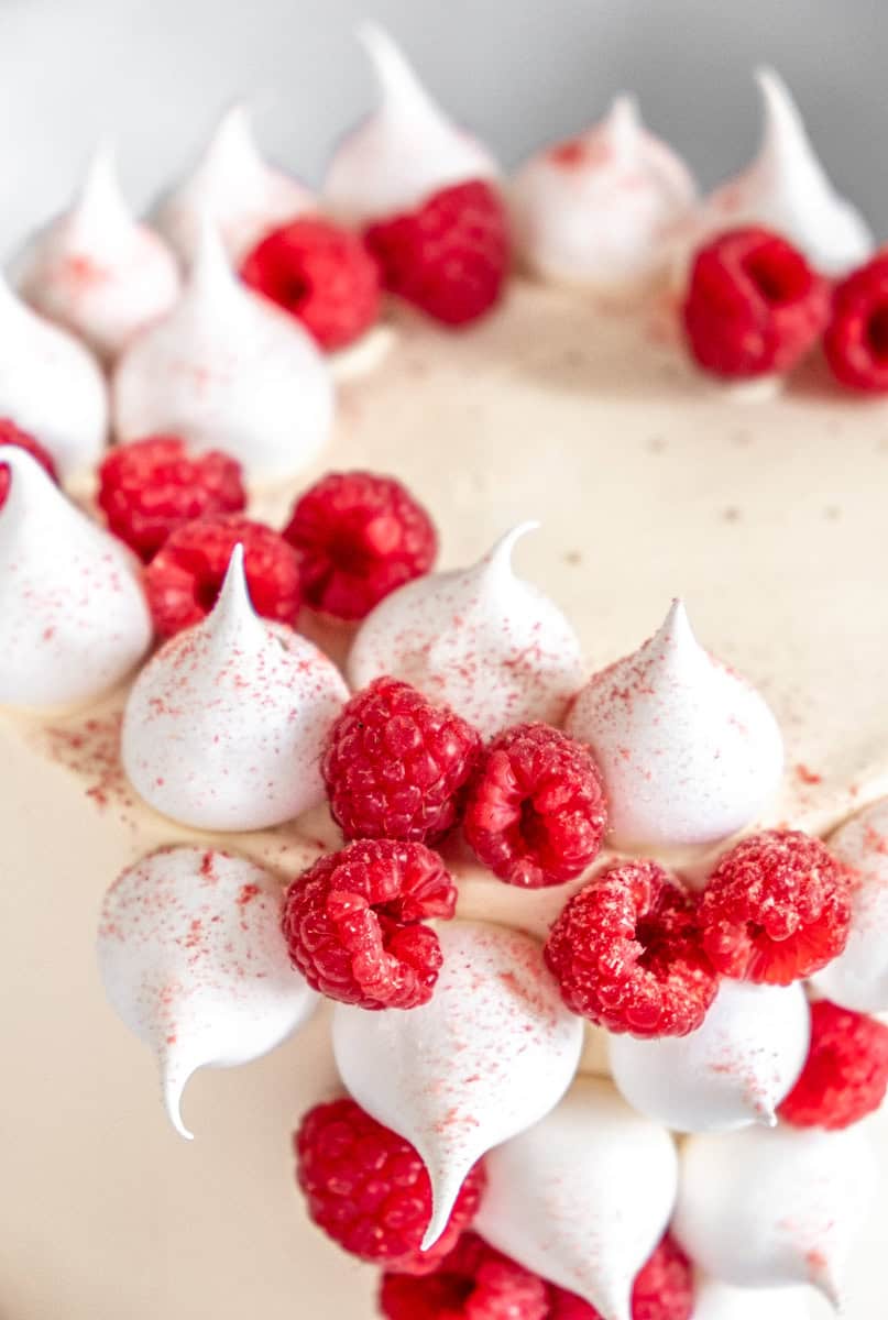 Mini Meringues shown decorating the sides of a cake next to raspberries. The meringues have been dusted with freeze dried raspberry powder.