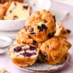 Images shows a few muffins stacked on a plate, one is sliced in half and you can see the blueberries inside.