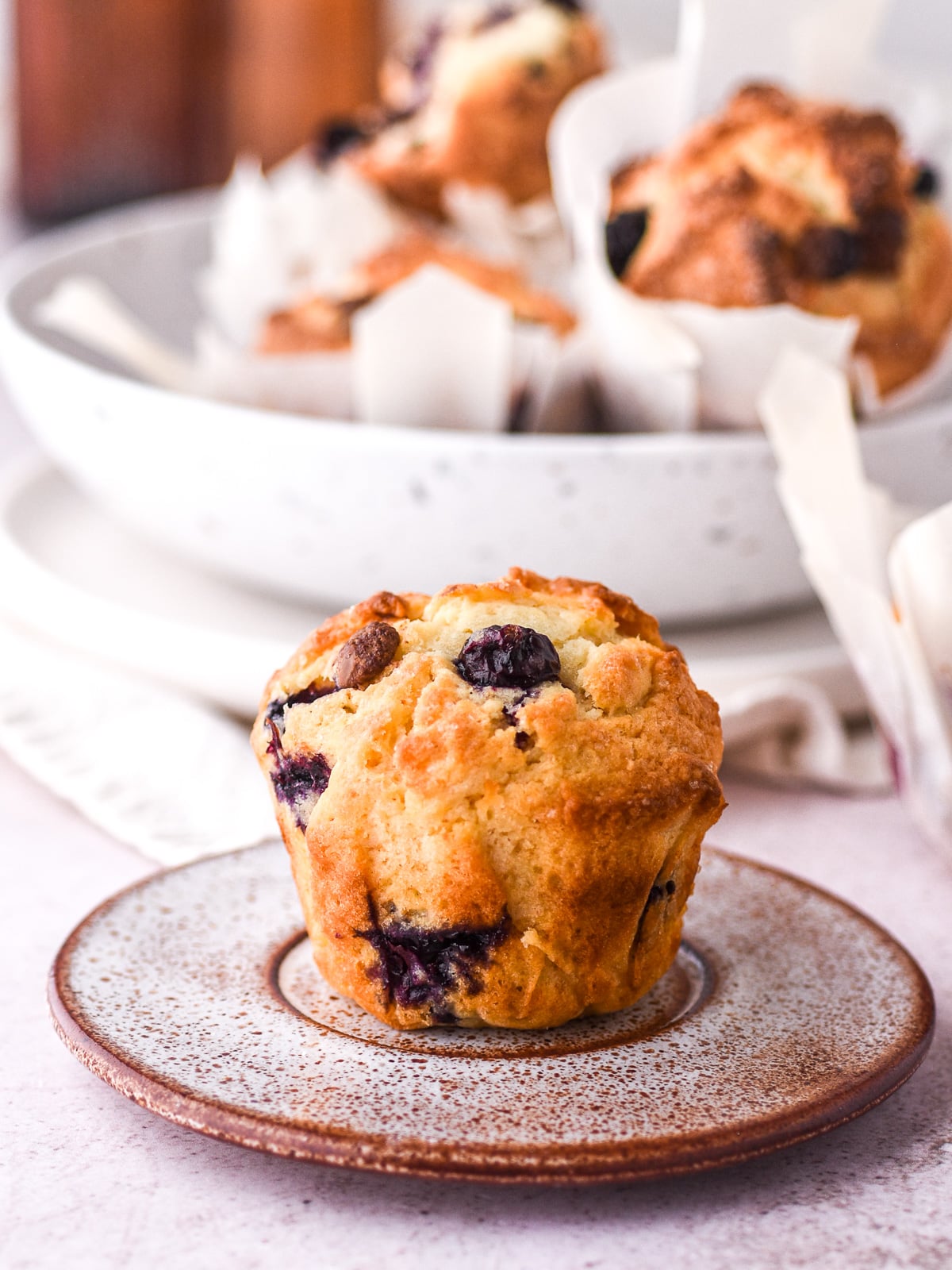 Image shows a single blueberry chocolate chip muffin on a plate, you can see a bowl filled with muffins in the background.