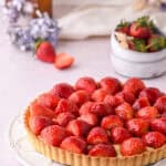 Strawberry Custard Tart shown on a white plate on a kitchen counter with flowers and strawberries in the background.