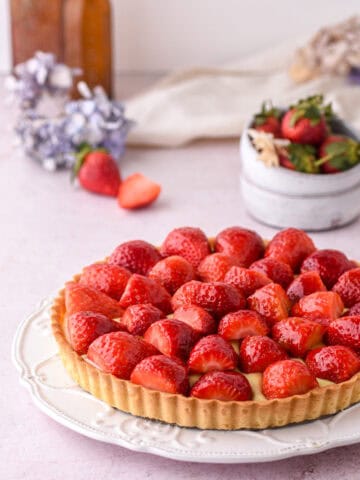 Strawberry Custard Tart shown on a white plate on a kitchen counter with flowers and strawberries in the background.
