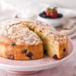 Berry cake shown on a pink cake stand, there are berries in a bowl in the background.