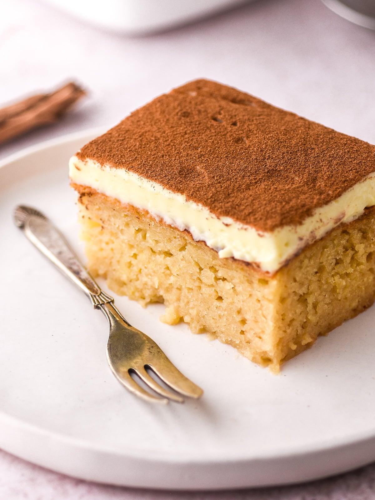 A slice of milktart malva pudding on a white plate next to a fork. You can see the moist layer of malva pudding and the creamy milktart layer.
