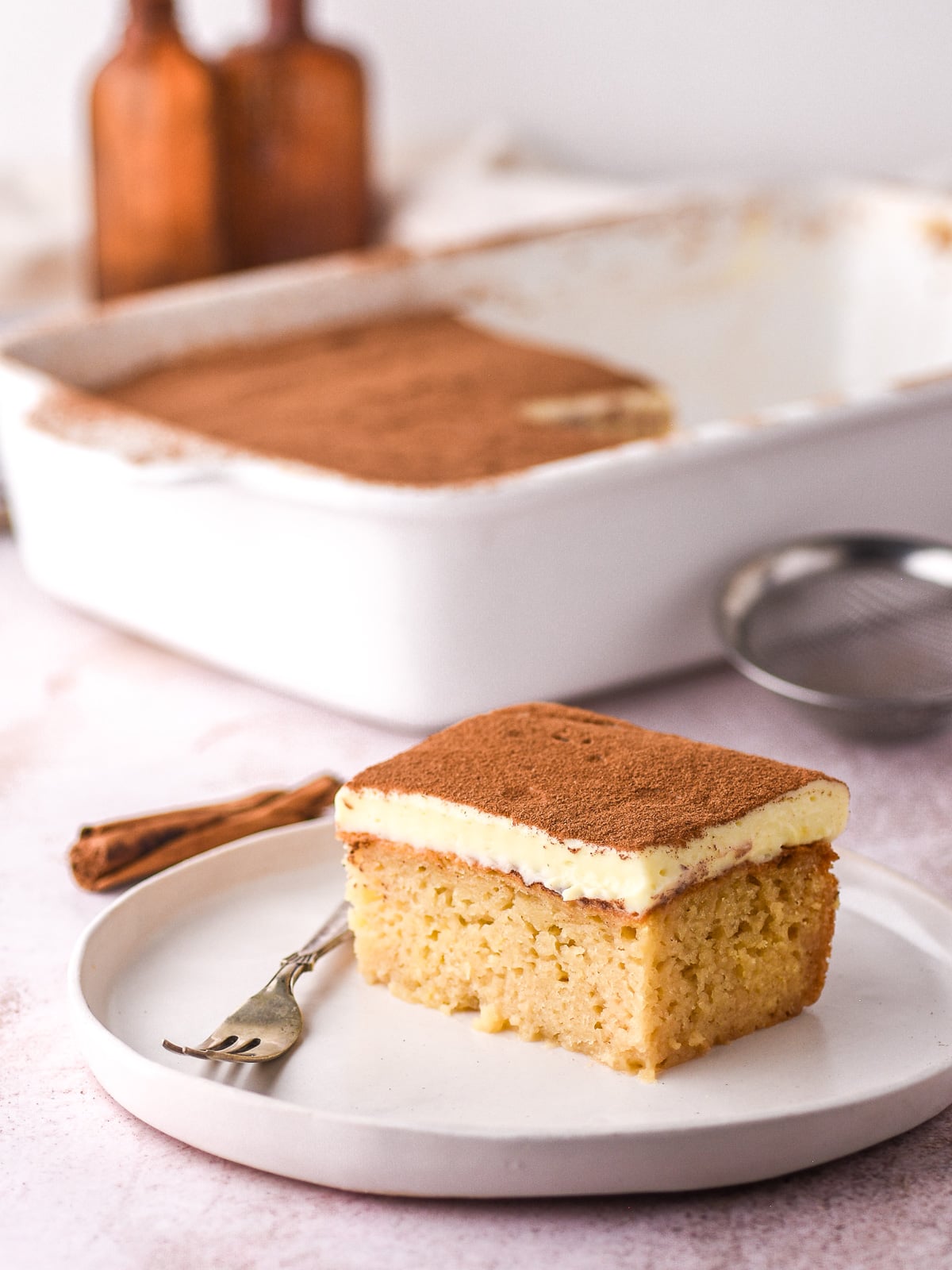 Image shows a slice of milktart malva pudding on a plate. In the background you can see the dish filled with the rest of the dessert.
