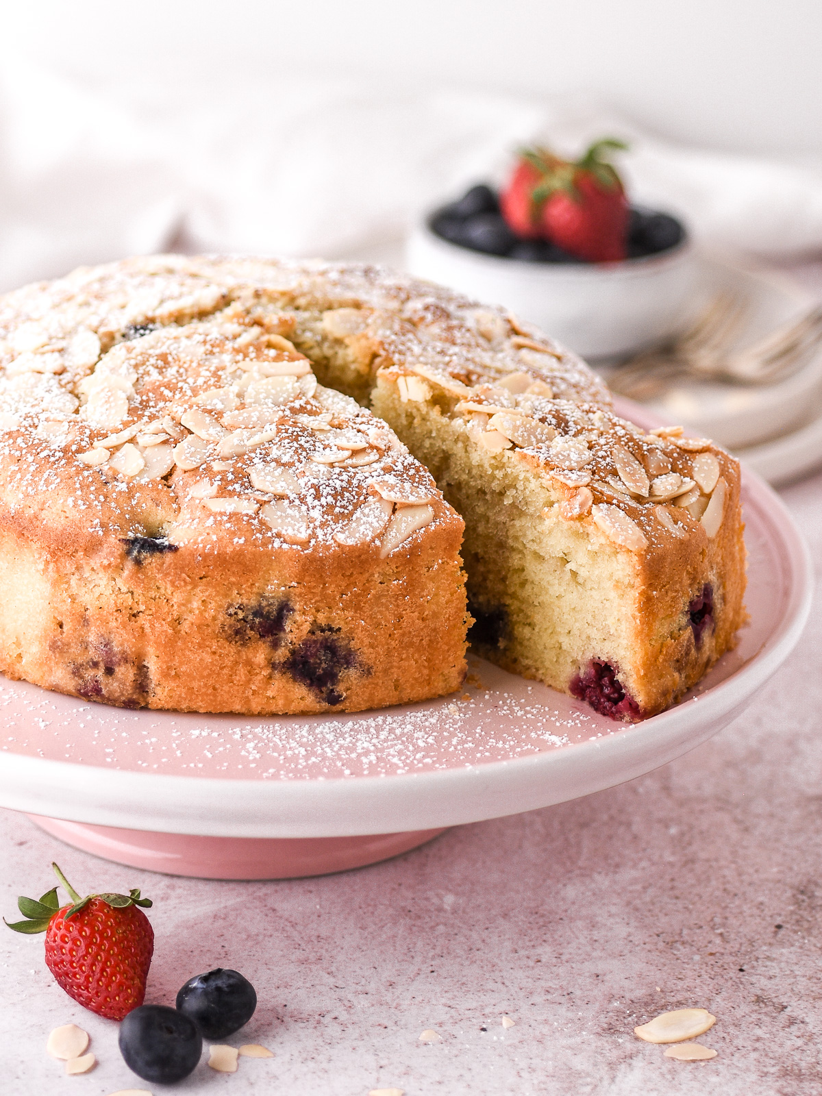 Image shows the whole mixed berry and lemon cake with one slice cut on a pink cake stand. There are blueberries and strawberries in the background and the foreground.