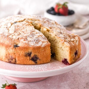 Lemon and mixed berry cake shown on a pink cake stand with one slice cut.