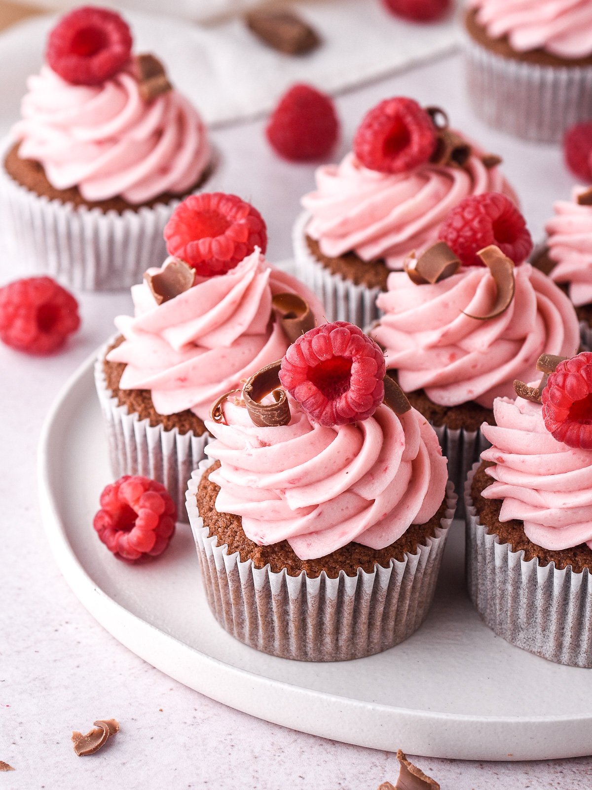 Raspberry chocolate cupcakes decorated with curls of chocolate and fresh raspberries shown on a white plate.