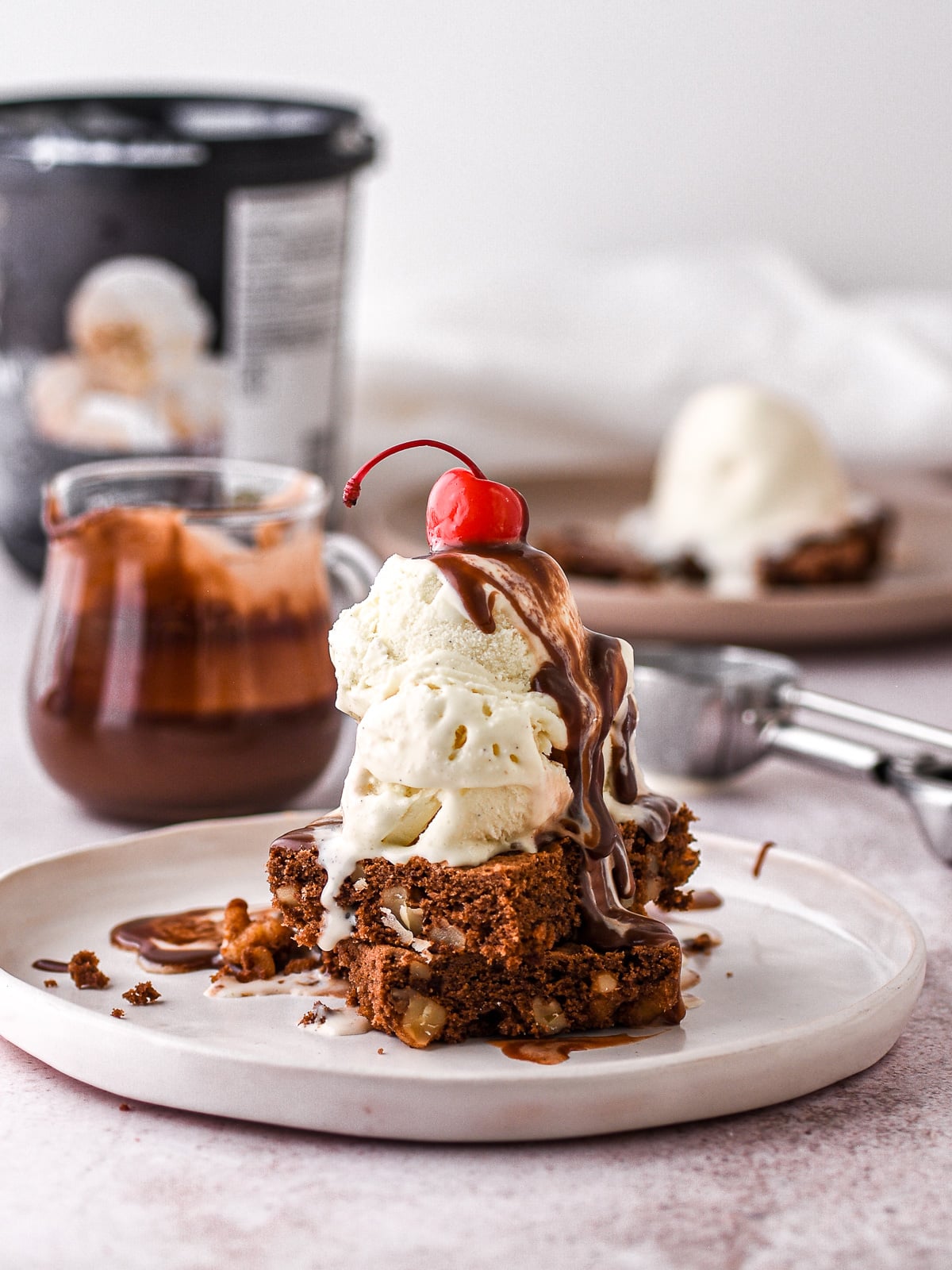 Image shows hot fudge brownie sundae, in the background there is a tub of ice cream and a glass jar filled with chocolate fudge sauce.