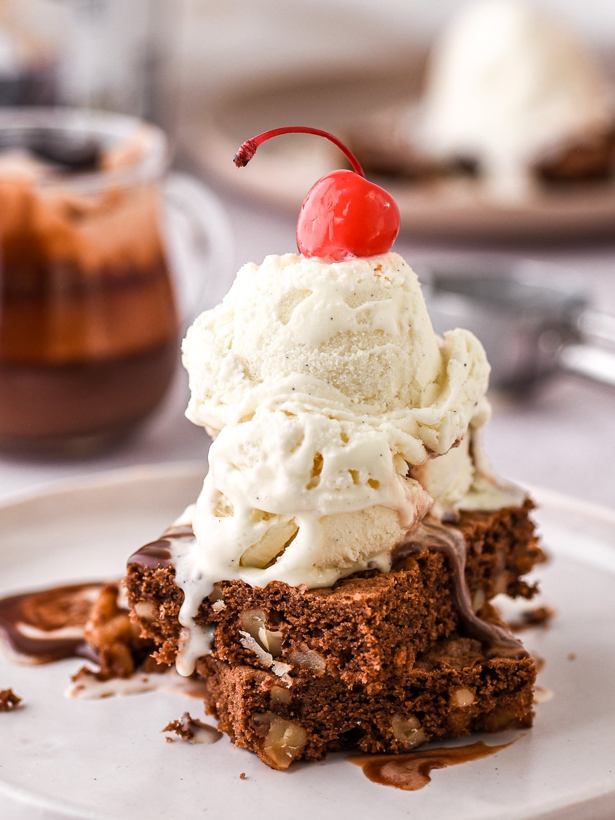 Image shows hot fudge brownie sundae, the brownie pieces are topped with vanilla ice cream, chocolate sauce, and a cherry.