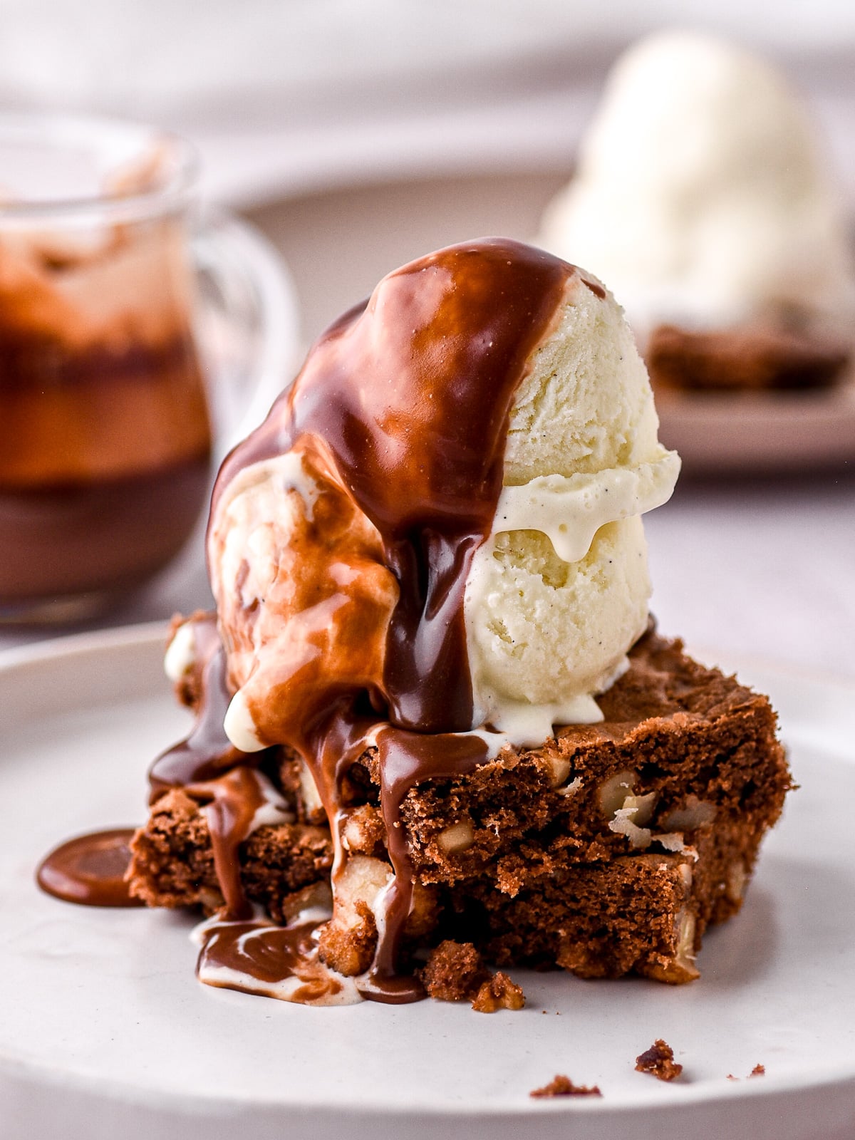 Image shows the hot fudge brownie sundae on a white plate. Two slices of walnut brownie topped with vanilla ice cream and covered in hot fudge chocolate sauce.