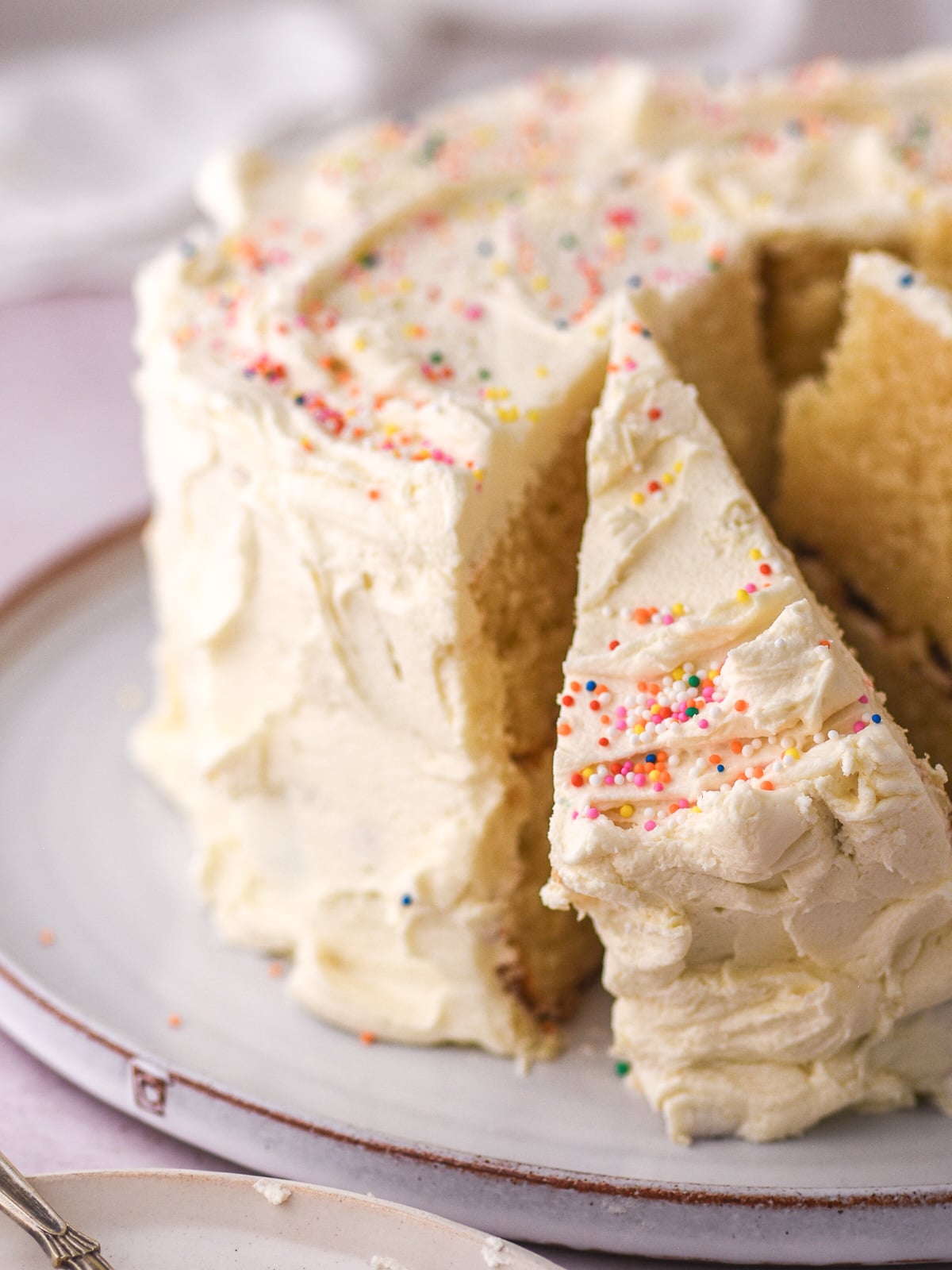 Image shows whole vanilla buttercream birthday cake on a cake plate, a few slices have been cut The cake is decorated with colourful sprinkles.