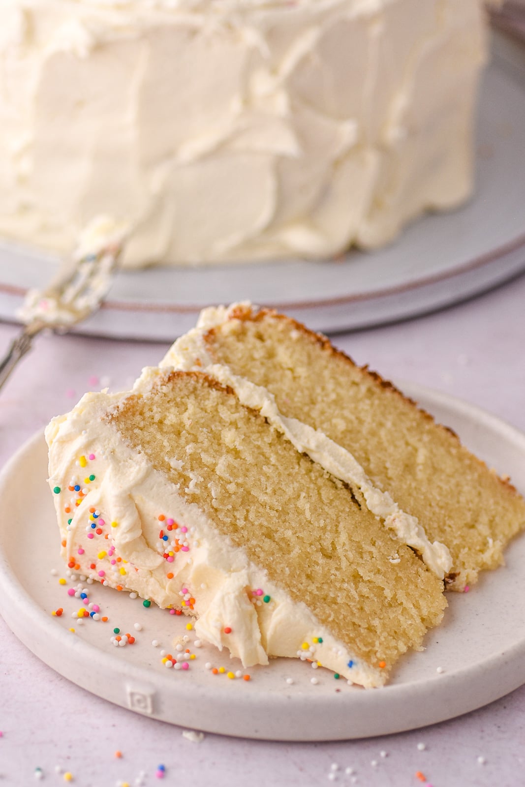 Image shows a slice of vanilla buttercream birthday cake on a white plate. There are sprinkles all around the cake.
