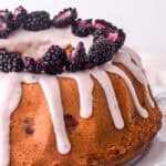 Cake shown on a white cake stand, you can see the lilac drip and fresh blackberries on top of the cake.