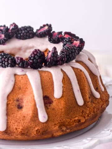 Cake shown on a white cake stand, you can see the lilac drip and fresh blackberries on top of the cake.