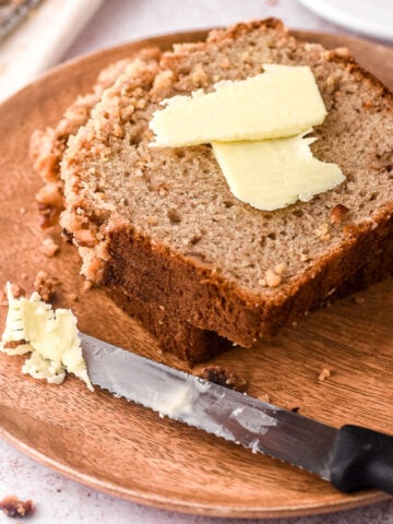 A slice of banana bread shown on a wooden plate with butter.