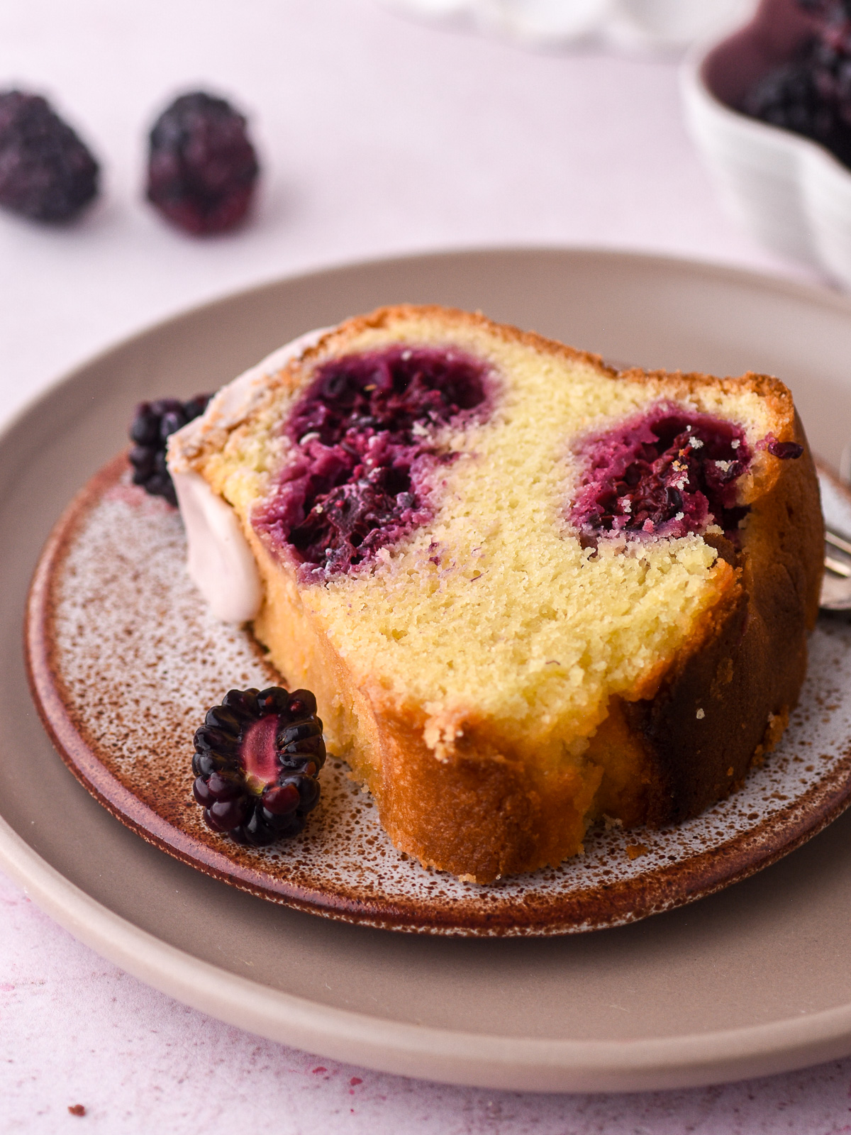 Image shows a slice of the cake on a small speckled cake, you can see the whole blackberries baked into the cake.