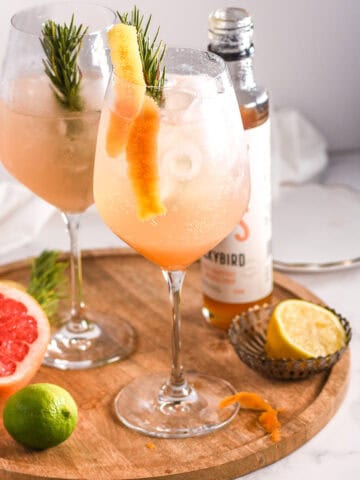 Gin spritz cocktail shown on a round wooden plate next to various citrus fruits.
