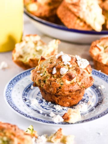 Zucchini and feta muffin shown on a blue plate, there are other muffins in the background.