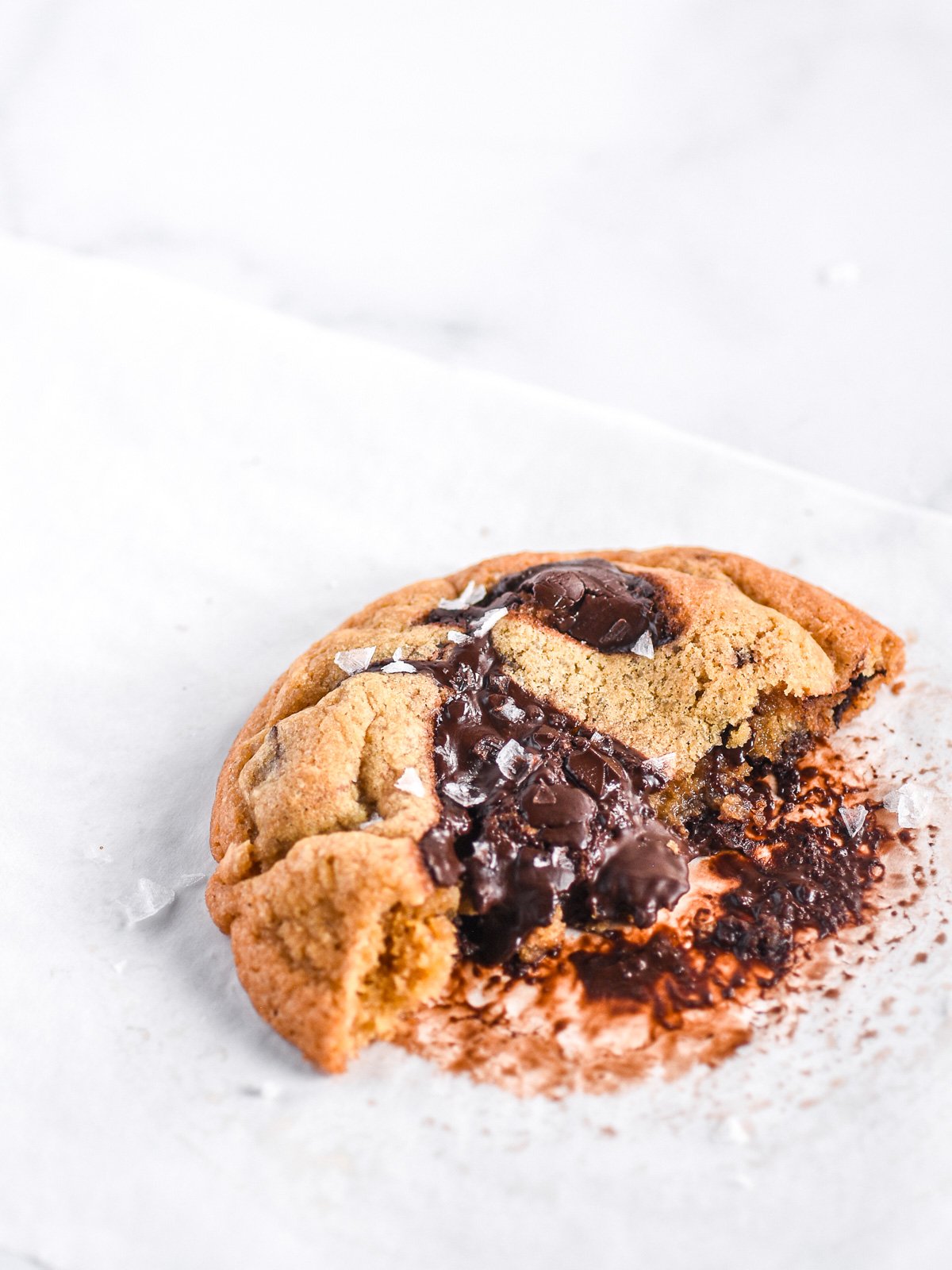 Half a cookie shown on a white surface, you can see the sea salt flakes and melted chocolate.