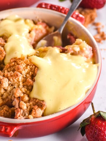 Strawberry & Apple Crumble shown in a red dish with custard.