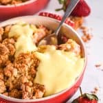 Crumble shown in a red ramekin, custard has been poured over the crumble.