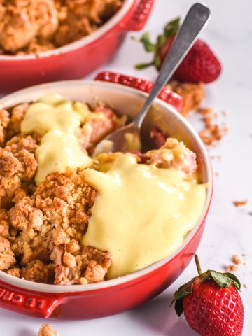 Crumble shown in a red ramekin, custard has been poured over the crumble.