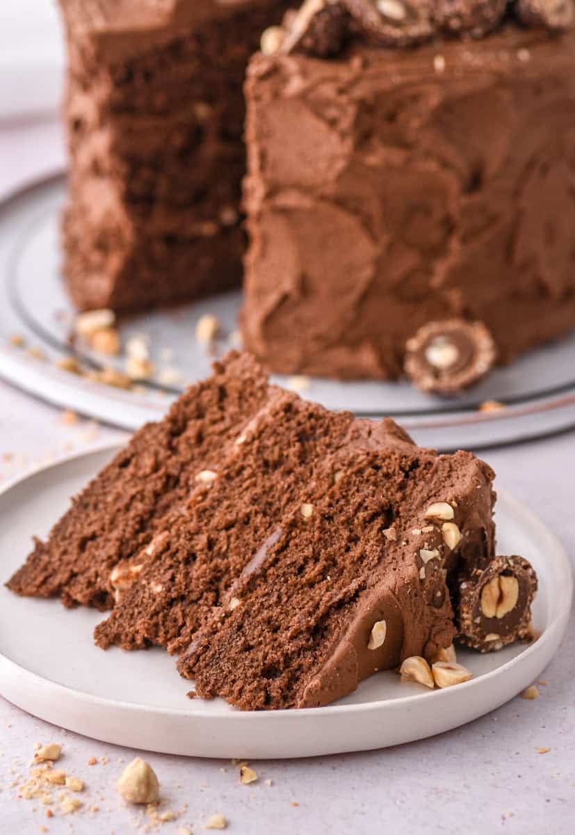 image shows a slice of cake on a white cake plate, the whole cake is in the background. 