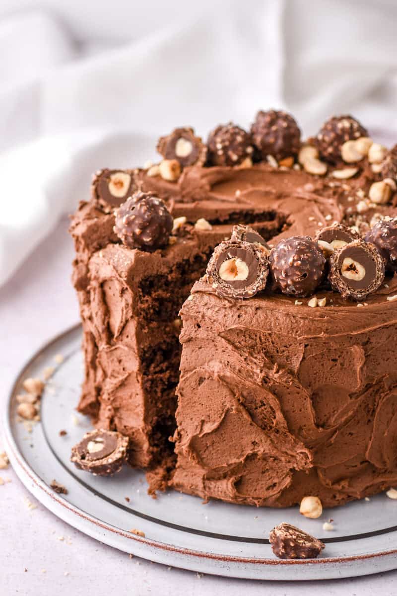 Image shows whole cake with a slice cut on a cake plate, the cake is decorated with Ferrero Rocher chocolate and chopped hazelnuts.