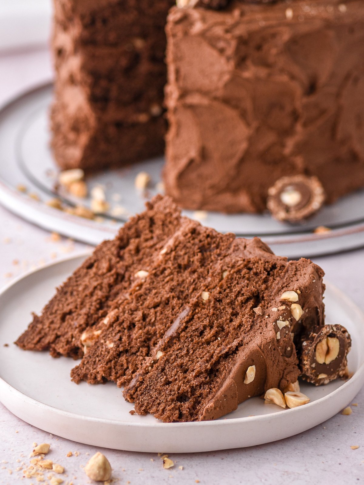 A slice of the Chocolate Nutella Ferrero Rocher Cake shown on a white plate with the whole cake in the background.